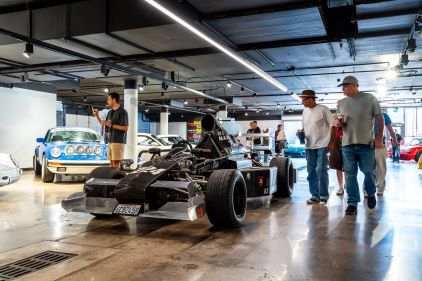 Visitors view a vintage Formula One race car displayed in a spacious showroom alongside other classic vehicles.