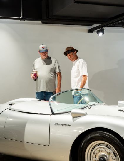 Two men in casual clothing stand beside a classic silver Porsche convertible.