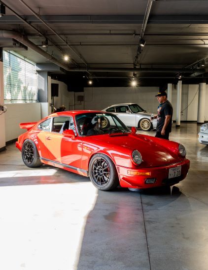 A red Porsche sits in a parking garage while a man in a blue shirt stands nearby observing it.