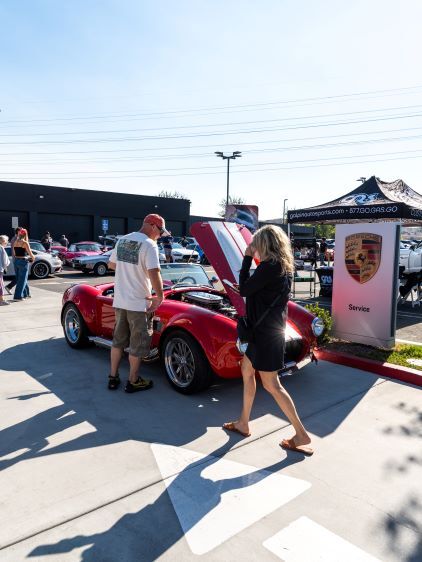 Two people examine the open hood of a red classic sports car at an outdoor car show.