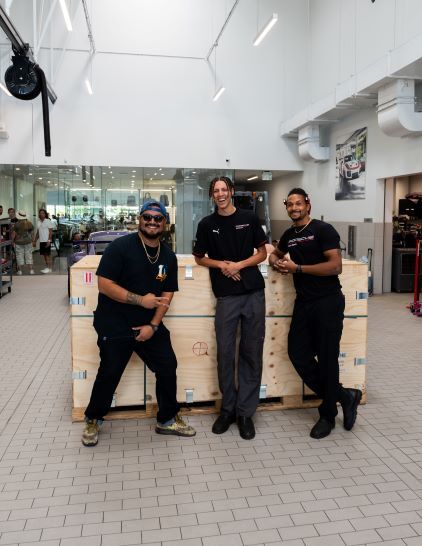 Three men in dark shirts pose next to a large wooden crate in a bright, modern building.