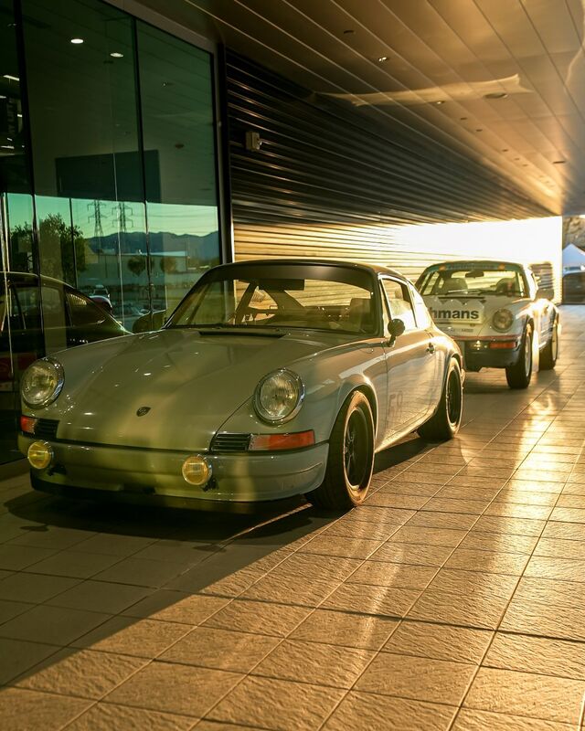 A vintage silver Porsche 911 sits parked in a sunlit driveway next to another classic car.