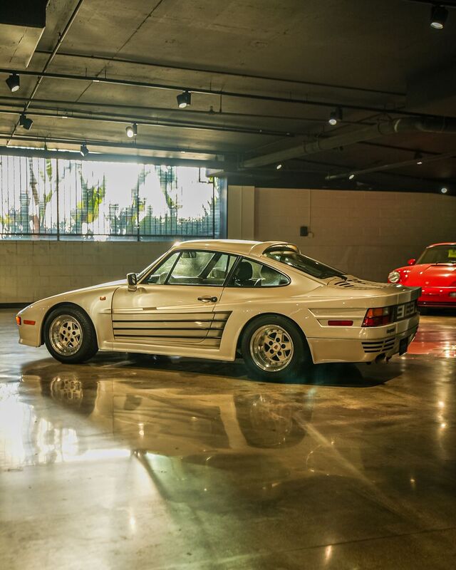A white Porsche 944 sits in a well-lit garage with a red car visible in the background.