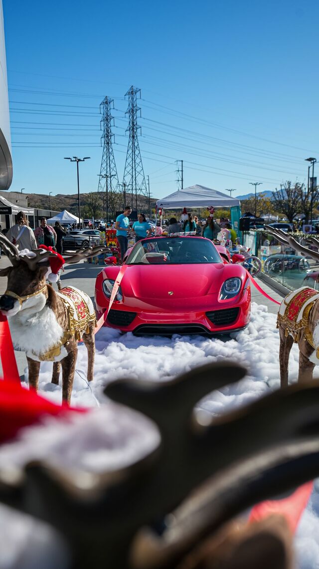A bright red Ferrari sits on a snowy display surrounded by reindeer statues at an outdoor event.