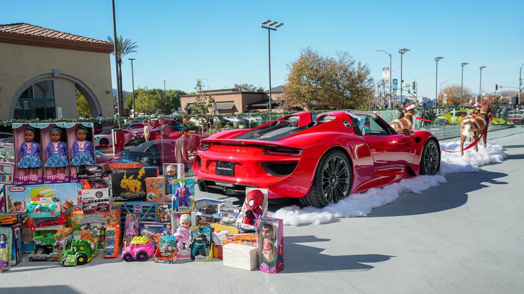 A red Porsche sits surrounded by toys and snow, with people dressed as reindeer nearby.