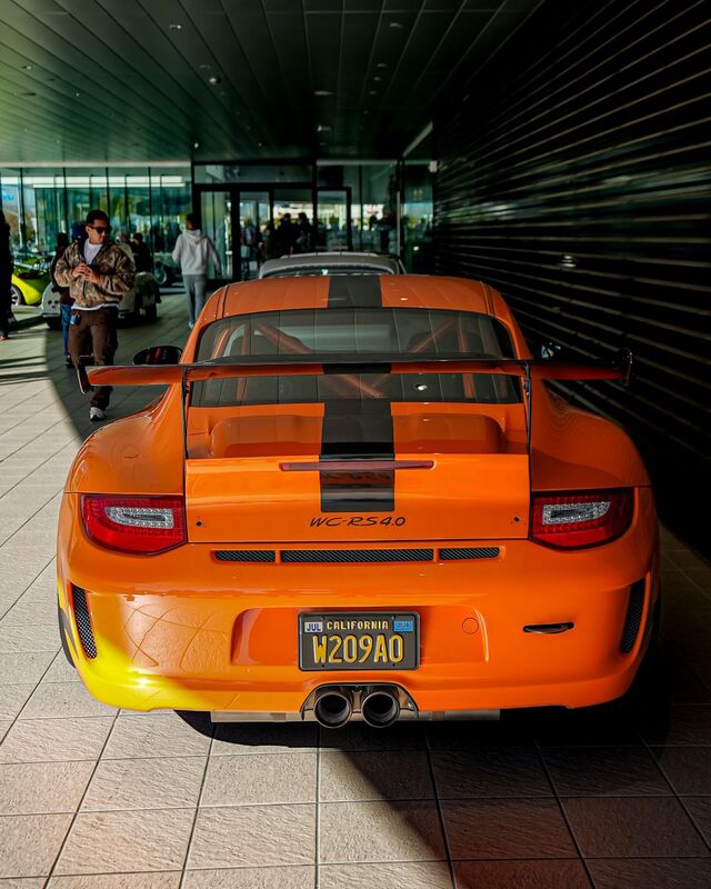 An orange Porsche 911 GT3 RS sits parked with its rear facing the camera near a building entrance.