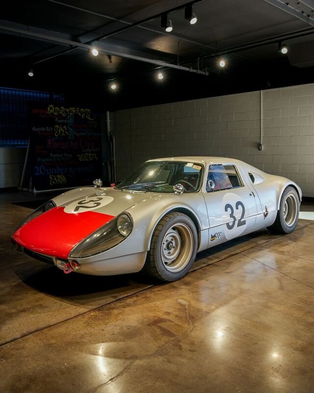 A vintage Porsche 32 race car sits in a garage with a chalkboard menu visible in the background.