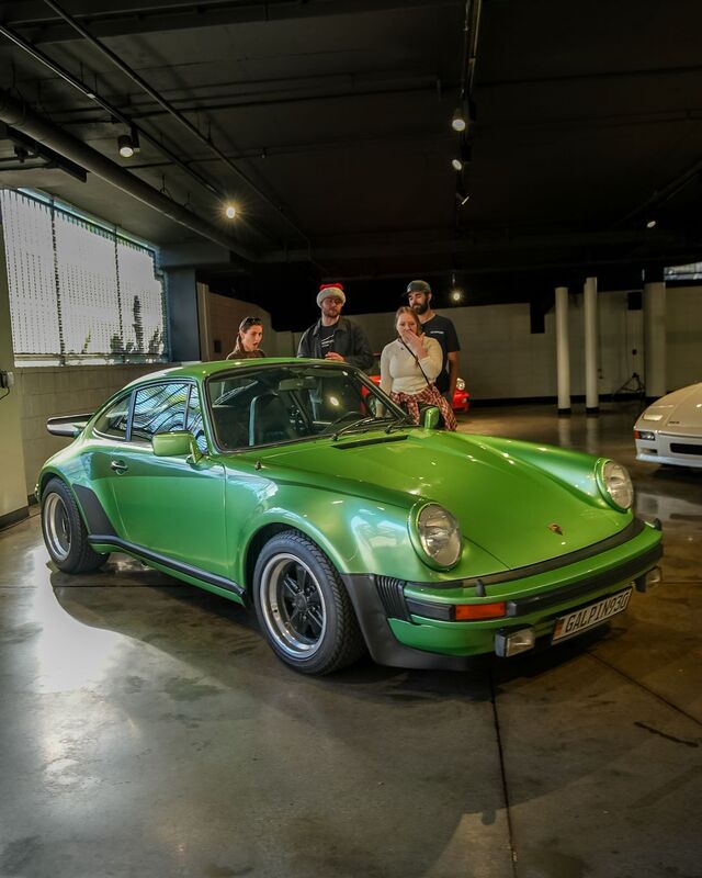 A bright green Porsche 911 sits in a garage as four people stand nearby, looking at the car.