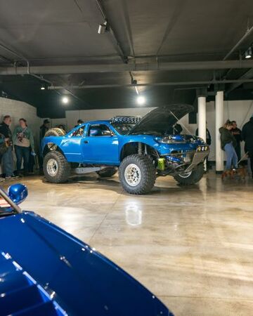 A bright blue off-road racing truck sits on display, with its hood open revealing the engine.