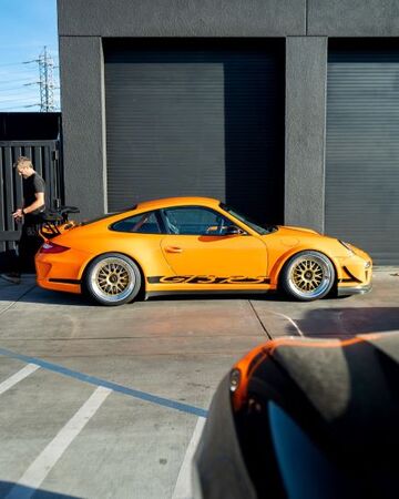 A vibrant orange Porsche is parked next to a man standing outside a modern building.