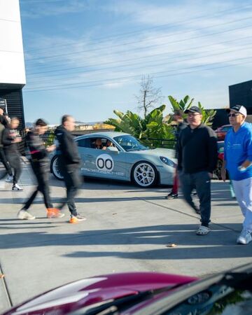 A gray Porsche car sits parked as a group of people in hats watch nearby.