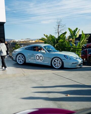 A silver Porsche 911 drives on a city street with tropical plants and a building in the background.