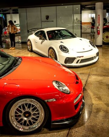 A woman stands near a white Porsche 911 GT3 inside a dealership, with a red Porsche parked in the foreground.