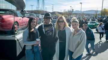 Four people pose for a photo at a car show with multiple vehicles and spectators in the background.