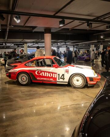 A red and white Porsche race car with Canon branding sits on display in a crowded showroom.