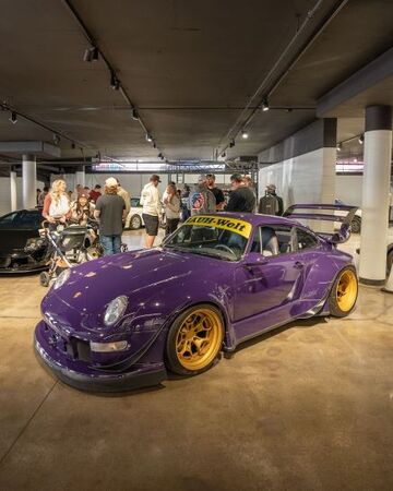 A purple Porsche 911 with gold wheels sits on display in a showroom surrounded by onlookers.