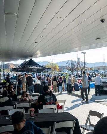 People gather at an outdoor event under a white metal canopy, with a small tent and vehicles visible beyond.