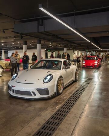 A white Porsche sports car is displayed in an indoor showroom surrounded by onlookers.