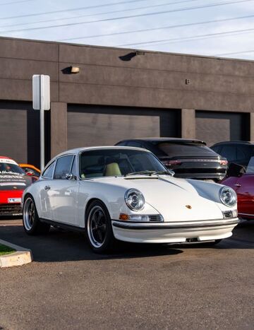 A classic white Porsche 911 coupe sits parked in front of a modern brick building.