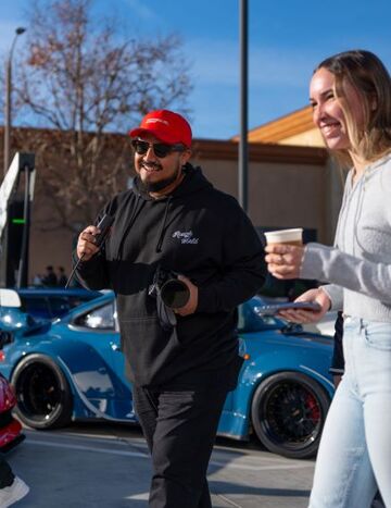 A man in a red hat and sunglasses takes a photo while a woman holds a drink and smiles nearby.