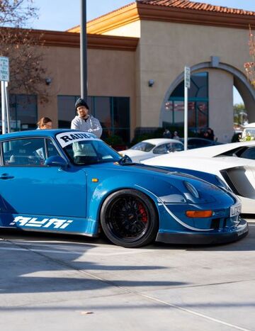 A man in a beanie leans against a vibrant blue classic Porsche at a car show.