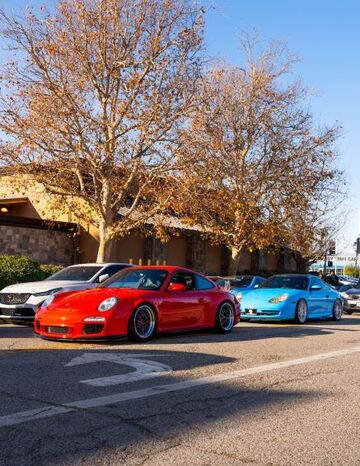 Several luxury sports cars, including a red Porsche, are parked along a street lined with trees.