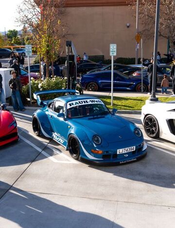 A blue Rauh-Welt Porsche 911 with a large wing sits in a parking lot surrounded by other cars.