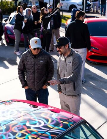 Two men in caps and jackets talk while gesturing near a colorful Porsche car, with a group of people nearby.