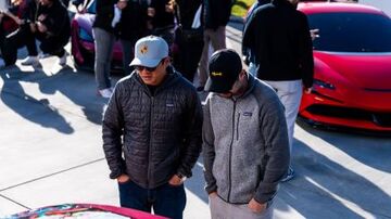 Two men in dark jackets and hats look intently at a bright red Tesla car.