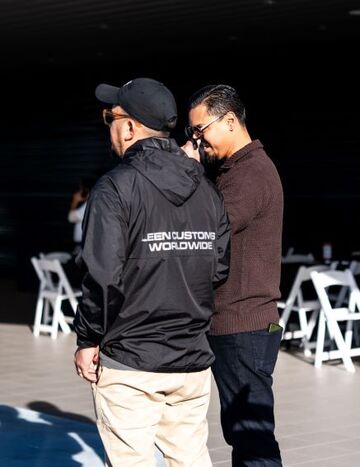 Two men stand discussing while a woman sits in the background at an event with white folding chairs.