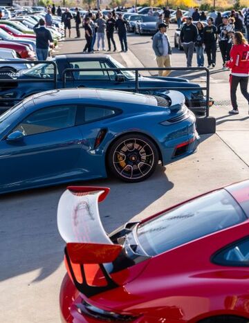 A blue sports car with a large spoiler sits in a parking lot surrounded by other cars and people.