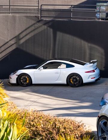 A white Porsche 911 coupe sits parked in a shadowy lot near greenery.