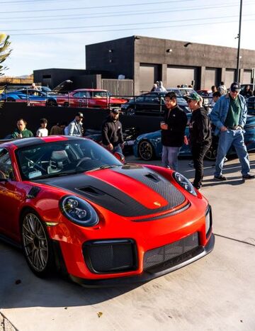 A vibrant red Porsche is showcased as a group of people admire and converse around it.