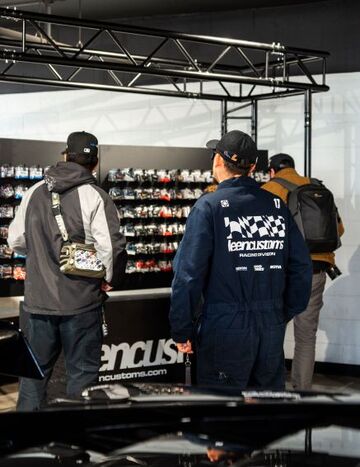 Three people in caps and jackets examine a display of custom wheels and automotive parts.