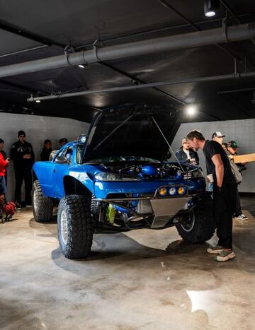 A bright blue lifted truck with its hood open is being examined by a group of people indoors.