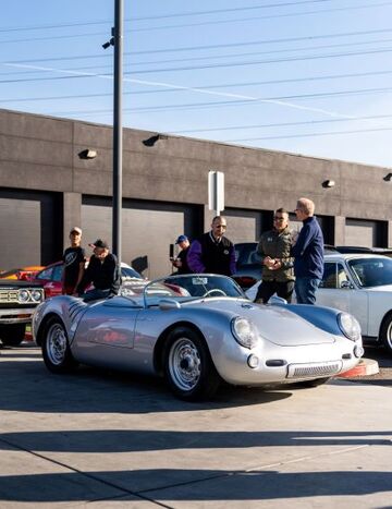 Several people stand near a vintage silver Porsche sports car during an outdoor event.