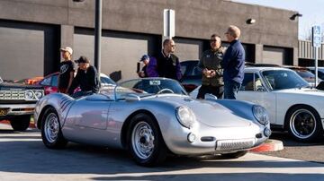 Several people examine a vintage silver Porsche Spyder parked among other classic cars.