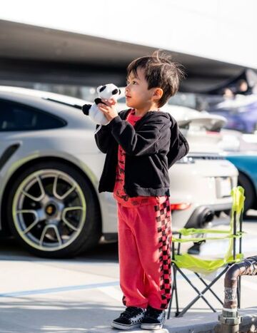 A young child in red and black checkered pants and a black hoodie holds a plush toy while standing near a parked car.