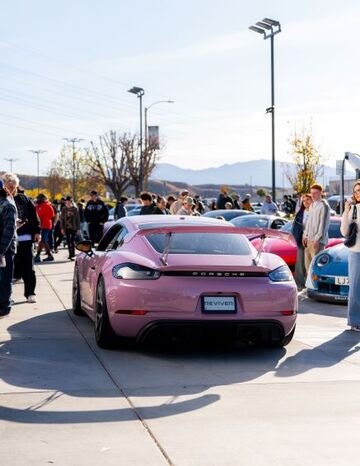 A pink Porsche sits on display as a crowd of people gather around it at an outdoor car show.