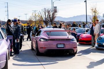 A light pink Porsche stands among a crowd of car enthusiasts gathered at a car meet.