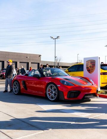 A red Porsche convertible sits at a car show, surrounded by other vehicles and onlookers.