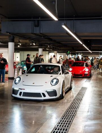 A white Porsche 911 is displayed in a car show alongside other luxury vehicles and a crowd of onlookers.