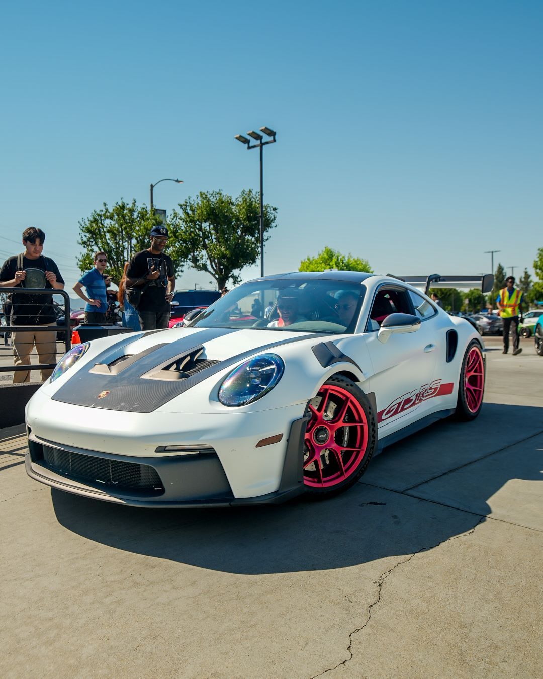 White Porsche 911 GT3 RS with black hood accents and bright pink wheels among people at an outdoor car show.
