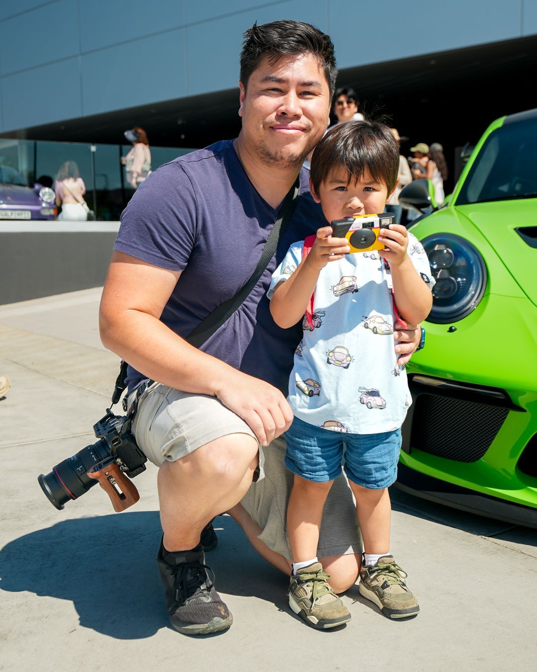 A man kneels beside a small child holding a toy camera, with a bright green sports car in the background.
