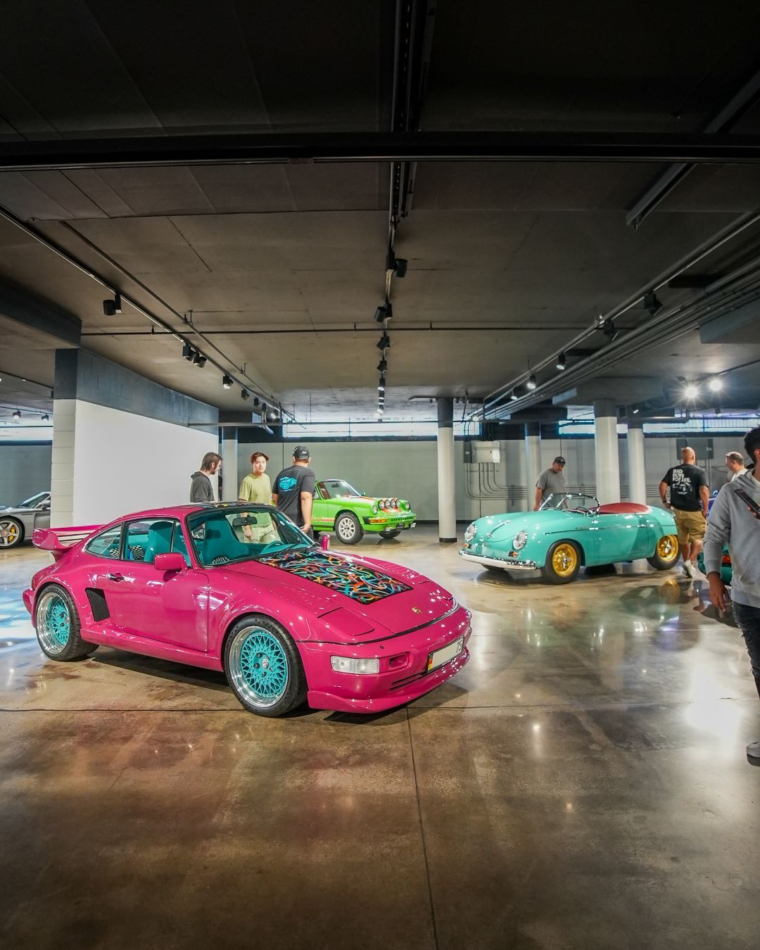 A bright pink Porsche with blue rims in a garage with other classic cars and people looking at them.