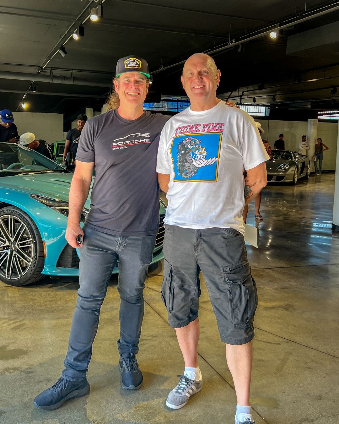 Two men smile for a photo at a car show, with a turquoise sports car behind them.