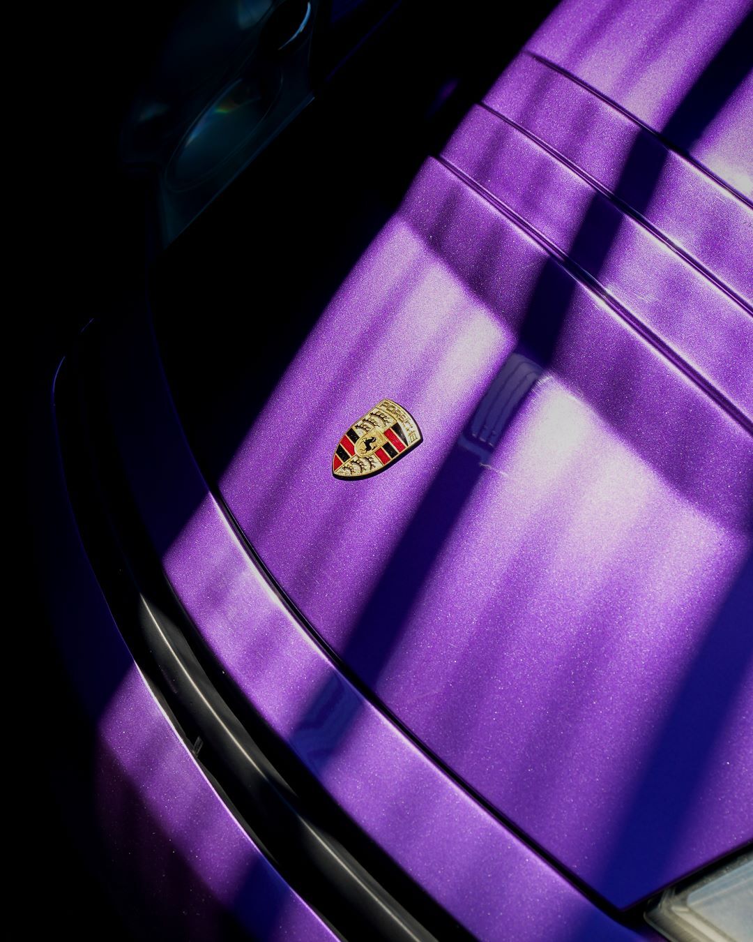 Close-up of a metallic purple Porsche car hood with its emblem, lit by diagonal sunlight and shadows.