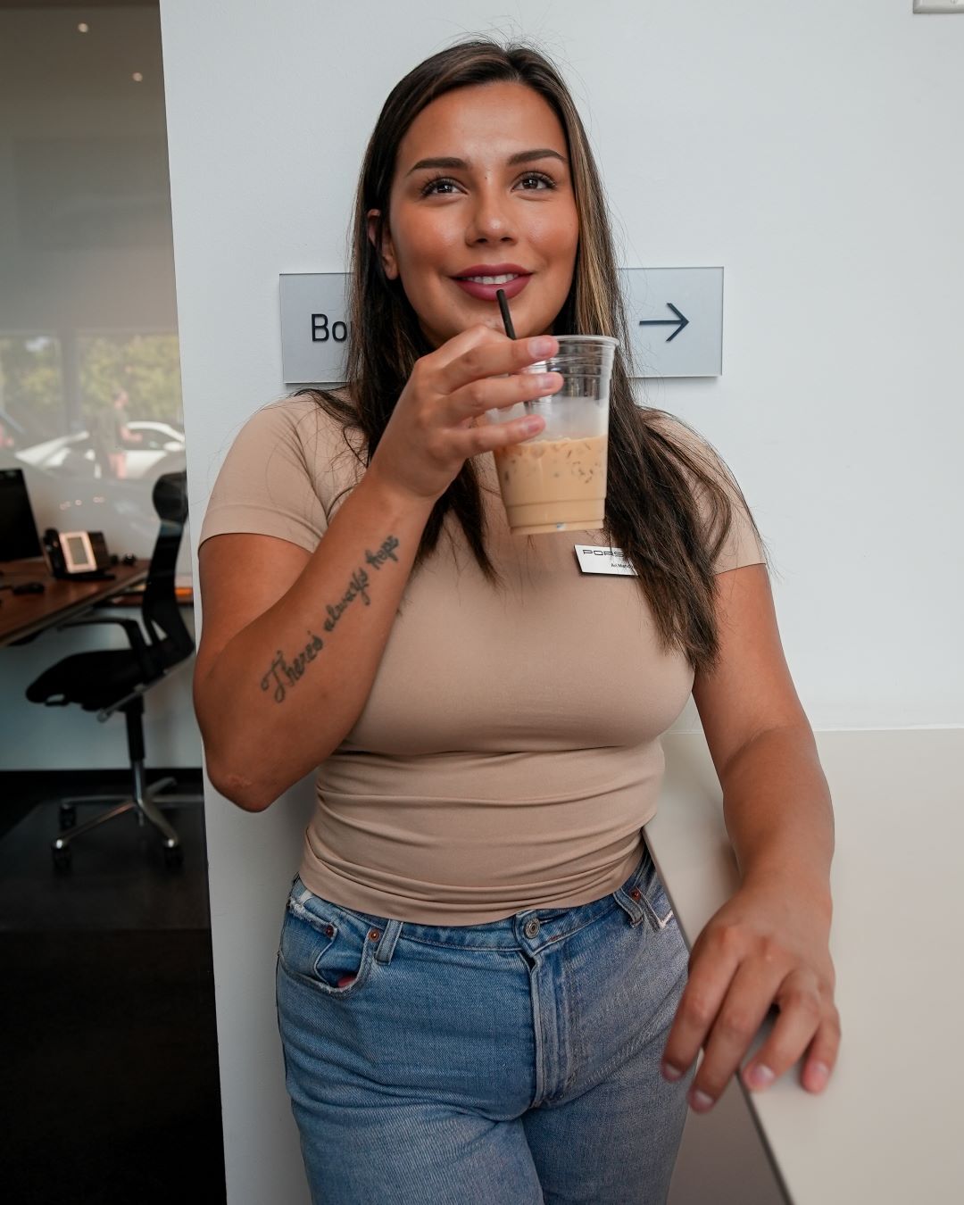 A smiling woman with long brown hair, wearing a tan shirt and jeans, sips an iced coffee from a clear cup.
