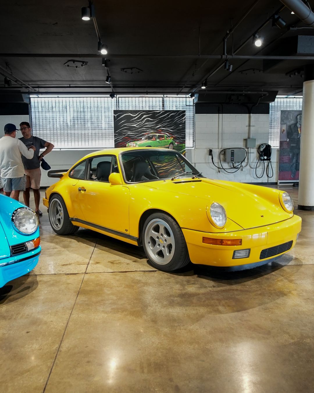 A vivid yellow Porsche 911, parked indoors on a concrete floor with people and artwork in the background.