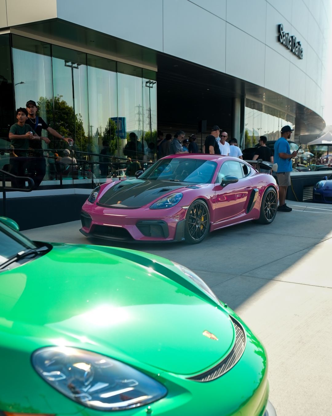 A green sports car's hood in the foreground with a magenta sports car behind, both parked outside a modern building.